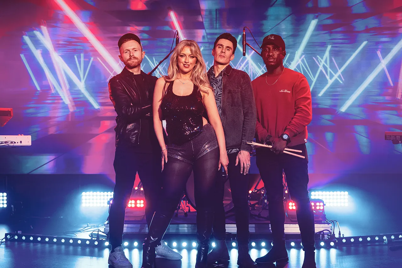 Four musicians posing on stage with colorful lights and laser beams in the background.